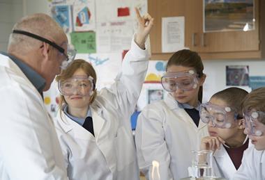 A teacher and younger high school students in a practical science class. The students are in uniform and they are all wearing lab coats and safety goggles. One girl is raising her hand.