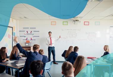 A male teacher stands at the front of a class pointing at equations on a whiteboard, while uniformed students listen and one raises their hand