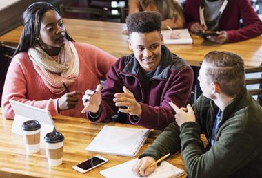 Three students sitting at a table with a tablet, pens, paper, having a discussion