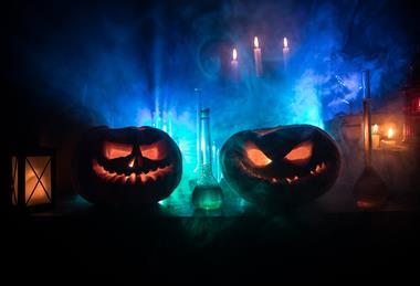 Two lit jack-o-lanterns sit on a table, surrounded by glass flasks and containers, candles and smoke, illuminated from behind by coloured lighting