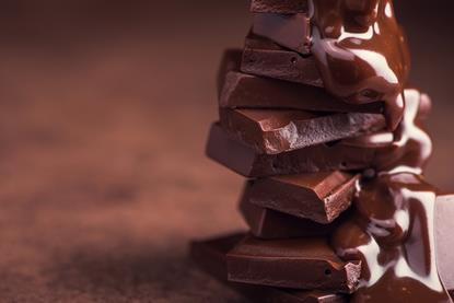 A close-up photograph of melted chocolate being poured over a pile of chocolate pieces against a light brown background