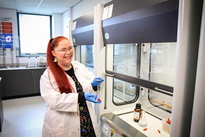 A happy teacher with a bold chemistry-theme-print dress and earrings wearing a lab coat whilst working at a fume cupboard