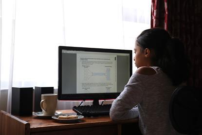 An image showing a girl studying a computer screen, sat at a desk, with a mug and biscuits to her left, all in front of a window