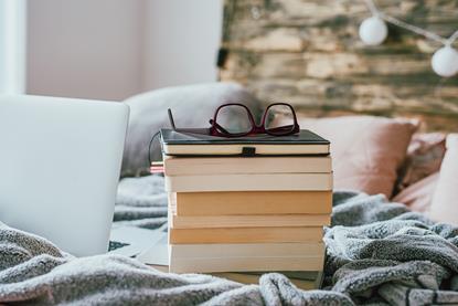 A stack of books on a bed with some reading glasses