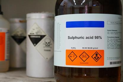 A close-up photograph of a bottle of sulfuric acid, with hazard symbols clearly visible on the label