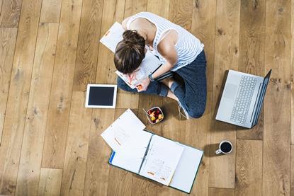 An image from above showing a girl sat cross legged, revising for exams, surrounded by a laptop, tablet, notes, coffee and snack