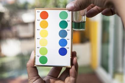 Hands holding a small glass container with water and universal indicator solution, comparing the green colour with a simple pH chart