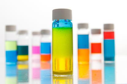A glass vial with a white lid containing a green liquid that is separating between blue at the top and yellow at the bottom. Other vials of different two-colour liquids are behind.