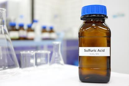 A small brown bottle labelled 'sulfuric acid' on a laboratory bench, with glass flasks and beakers in the background
