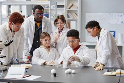 Group of children in lab coats and safety glasses doing a practical with their teacher looking on