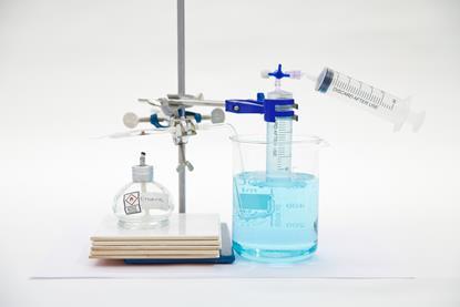 An ethanol burner sits on tiles with a pipette above while a clamp holds an inverted syringe in a beaker. A tube goes from the pipette into the liquid in the beaker. A second syringe is attached to the first with a 3-way Luer connector