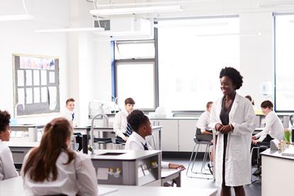 A female teacher stands at the front of a school laboratory wearing a white lab coat, smiling and speaking to students seated at their desks wearing uniform