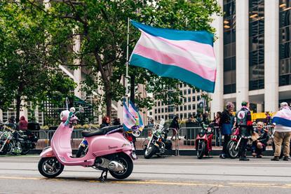 A pink moped and person carry a trans pride flag on a San Francisco street