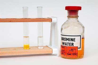 A bottle of bromine water next to two test tubes - one contains only clear liquid and the other contains clear liquid sitting on an orange liquid