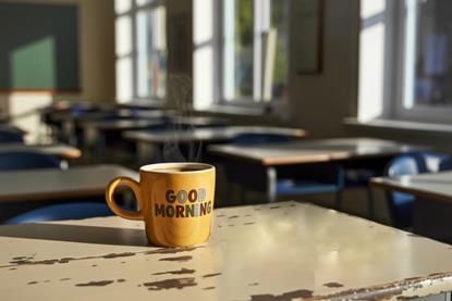 A colourful mug which says Good Morning with steaming coffee on a teacher's desk at the front of an empty classroom before the school day begins.