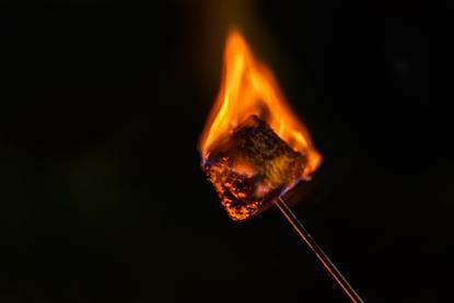 A close-up photograph of a burning marshmallow on a metal skewer, pictured against a dark background