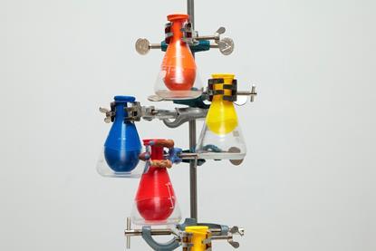 A number of glass conical flasks attached by clamps to a metal stand. Each flask has a colourful balloon sealed around its neck that has been sucked inside the flask.
