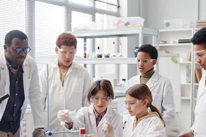 A teacher and a group of students are in a lab. Two of the students are carrying out a practical activity by adding one solution to another while the other students and the teacher observes them.