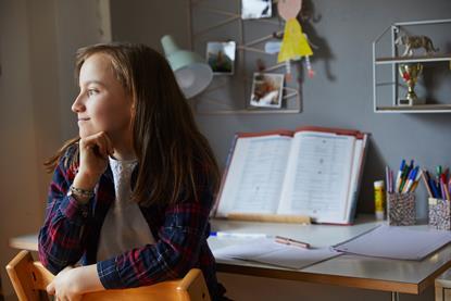A picture showing a girl sitting on a chair and gazing out of the image, behind her is a desk with lamp, books and pens