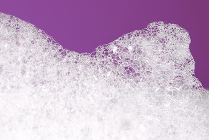A macro photograph of a foamy lather from soap or detergent against a plain purple background.