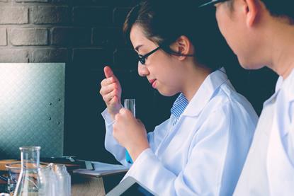 A photograph showing two people in lab coats; the person in focus is carefully smelling the contents of a test tube
