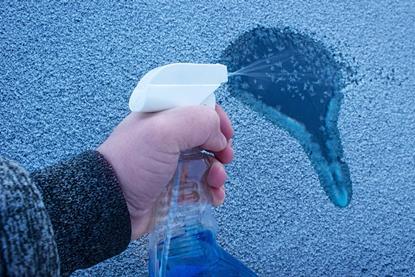 A hand spraying blue liquid on a window covered in ice which is melting