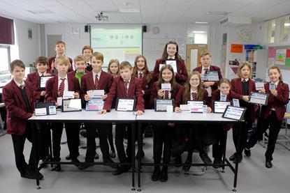 A group of secondary school students standing around a table in a classroom. The students are the front of the group are holding tablets displaying the home page of the I'm a Scientist, Get me out of here website.