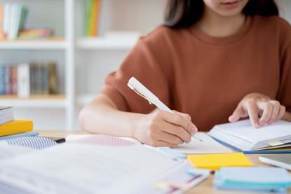 A student sitting at a desk, reading from a book while making notes