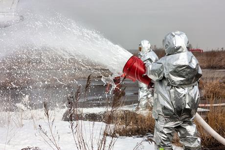 Firefighters in protective silver suits spray a thick white foam from a large hose