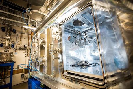 A technician in a white cleansuit makes adjustments inside a large piece of metal scientific equipment