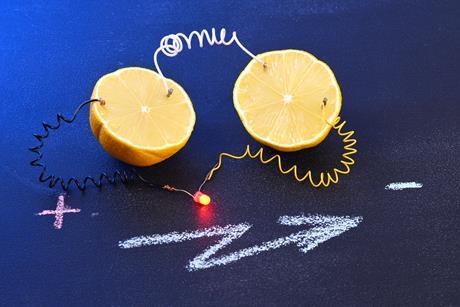 Two halves of a lemon wired together and to a red bulb that is glowing. On the table written in chalk is a plus and minus sign and a lightning bolt