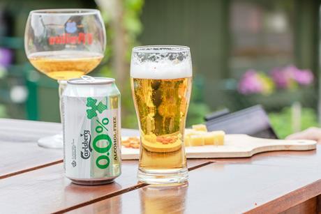A can of Carlsberg 0.0% alcohol-free beer and a glass sitting on a picnic table outdoors with some snacks on a sunny day