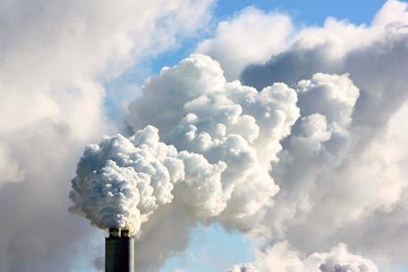 A photograph of the top of a factory chimney shown against the sky, emitting a dense smoke into the air