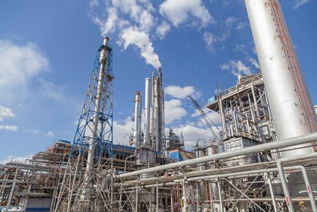 A fractional distillation tower as part of a large industrial plant with lots of pipes, scaffolding and chimneys giving off emissions.