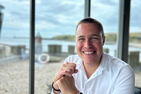 Adam Savin is smiling at the camera, wearing a white shirt. The sea is in the background.