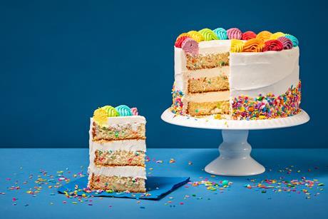 A cake on a stand with a piece taken out on a paper napkin. The cake is colourfully decorated and has sprinkles baked into the batter.