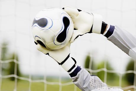 A football goalkeeper wearing protective gloves stretching to block a football from going into the goal