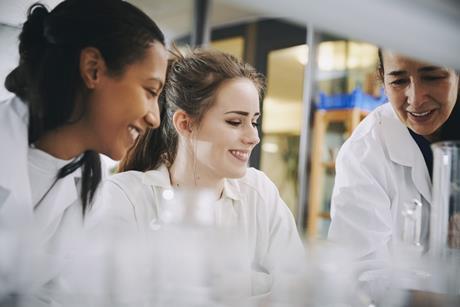 A science teacher works with two students at a laboratory bench; all three people are smiling, looking at some experimental apparatus