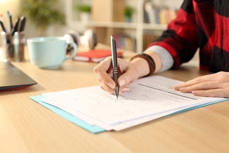 A student's hands holding a pen and writing on an exam paper on a desk; a notebook, mug and laptop are visible in the background