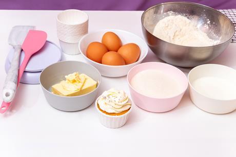 A finished cupcake with the ingredients and tools to make cupcakes including eggs, butter flour, sugar, scales, bowls, paper cups and spatulars