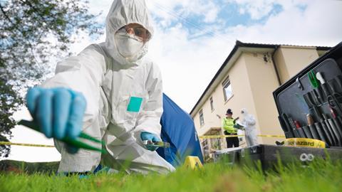 A crime scene technician wearing white overalls and latex gloves collects evidence from a lawn with some tweezers