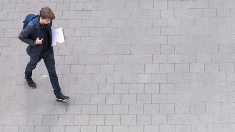 A top view of a student dressed in blue carrying a rucksack walking across grey paving stones, from left to right