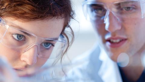 Two students wearing safety goggles and lab coats closely observe a reaction