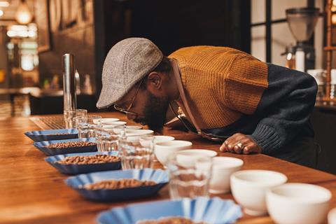 An image showing a man wearing a flat cap and glasses behind a table covered with bowls of different kinds of coffee beans, glasses and cups. He's testing the quality of the coffee beans by smelling them.