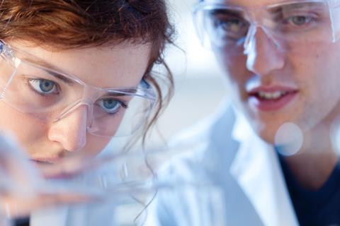 Two students wearing safety glasses and lab coats closely observe a reaction
