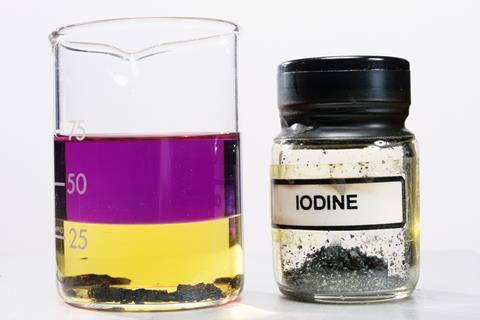 A glass lab beaker with a liquid in that is purple at the top and yellow at the bottom with a black substance at the bottom. Next to is is a jar labelled iodine containing more black substance