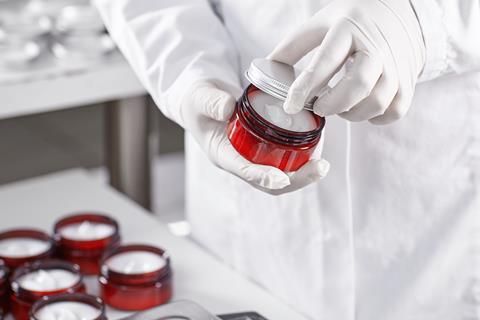 A scientist in a white coat and gloves opens a jar of cosmetics