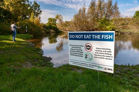 A sign by a lake warning against eating fish from the lake due to high levels of PFAS. In background is a man fishing with a rod.
