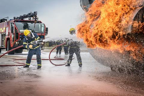 Two firefighters tackling a blaze on an aircraft with water hoses and the fir engine in the background