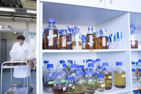 An image showing chemicals in bottles in a cabinet; a technician can be seen in the background pushing a trolley of glassware